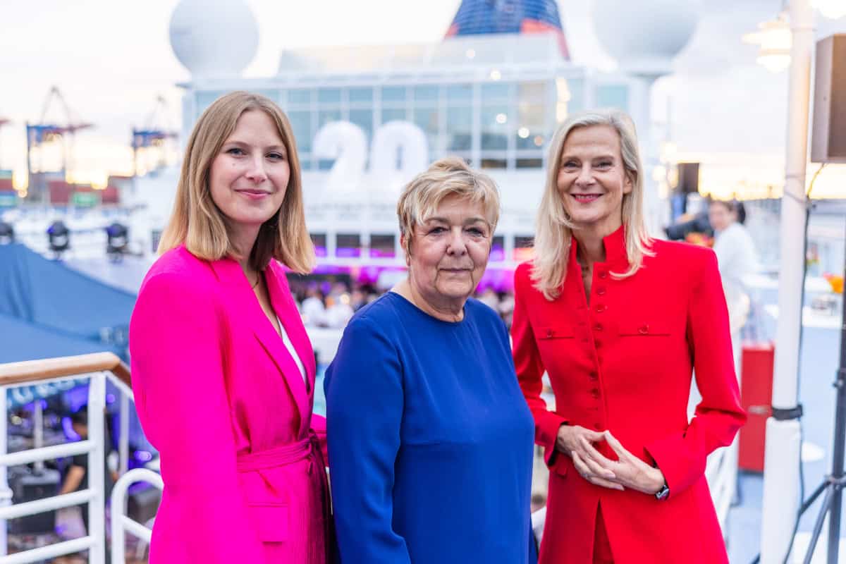Drei Frauen stehen nebeneinander auf dem Deck eines Kreuzfahrtschiffes. Sie tragen farbenfrohe Blazer in Pink, Blau und Rot und lächeln in die Kamera.