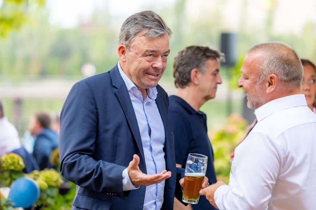 Ein Mann in einem dunkelblauen Sakko spricht mit einem Mann in einem weißen Hemd, der ein Glas Bier hält, während weitere Personen im Hintergrund stehen.