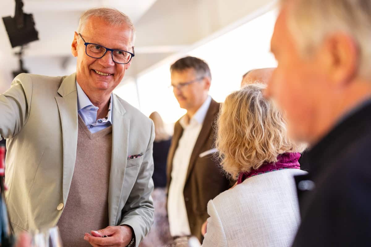 Ein lächelnder Mann mit Brille und beigem Sakko steht im Vordergrund, während im Hintergrund weitere Personen in einer hellen Räumlichkeit zu sehen sind.