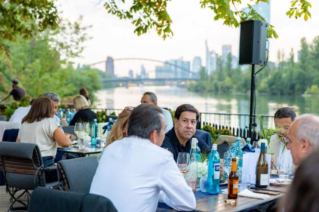 Eine Gruppe von Menschen sitzt an gedeckten Tischen auf einer Terrasse am Flussufer, im Hintergrund ist die Skyline einer Stadt mit einer Brücke zu sehen.