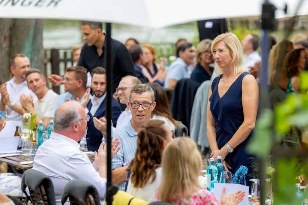 Eine blonde Frau in einem dunkelblauen Kleid steht bei einer Outdoor-Veranstaltung an einem Tisch, während Gäste im Hintergrund applaudieren und sitzen.
