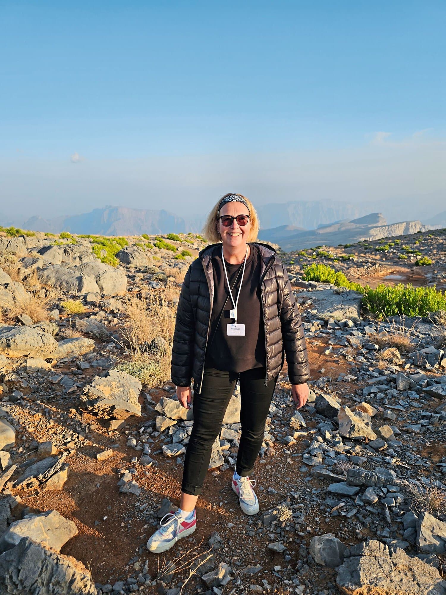 Eine Frau mit blondem Haar, Sonnenbrille und schwarzer Steppjacke steht auf einem felsigen Bergplateau vor einer weiten, bergigen Landschaft bei Tageslicht.