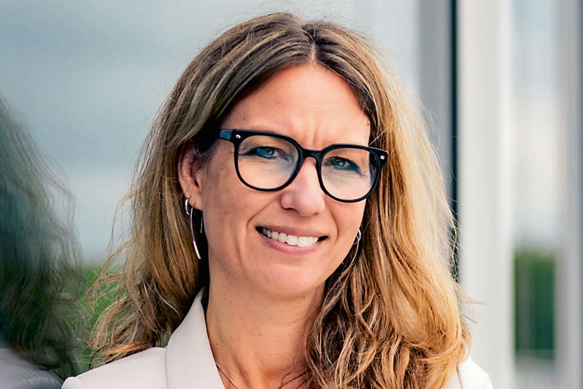 Eine Frau mit langem, hellbraunem Haar und einer schwarzen Brille lächelt freundlich in die Kamera. Sie trägt eine helle Jacke vor einem hellen Hintergrund.