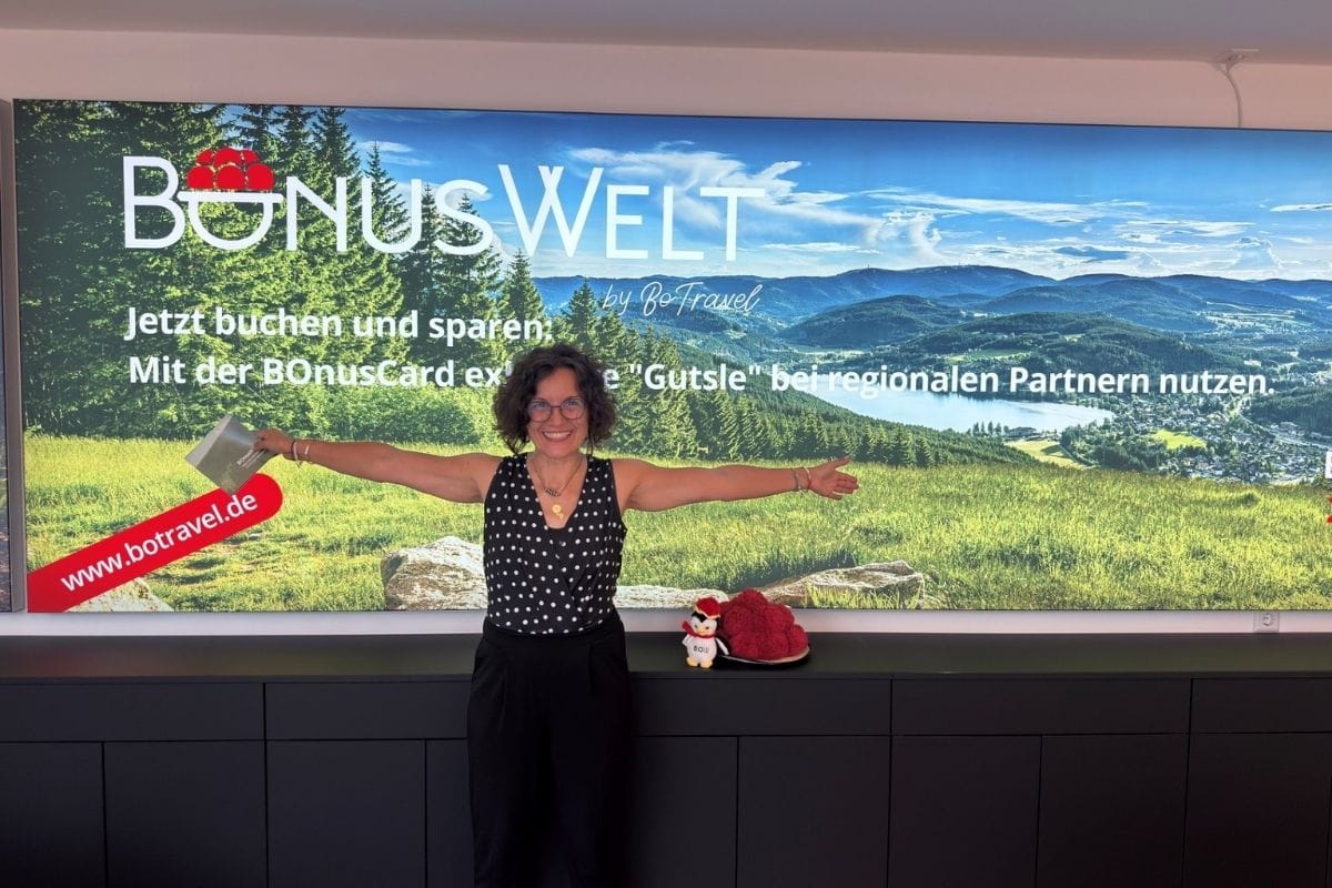 Eine Frau mit Brille steht vor einer großen Werbewand mit Landschaftsmotiv, hält ein Heft in der Hand und hat die Arme weit zur Seite ausgebreitet.