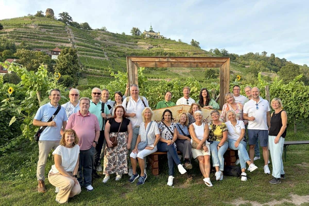 Eine große Gruppe von Menschen posiert vor einem hölzernen Bilderrahmen in einem Weinberg, im Hintergrund sind ein Schloss und bewirtschaftete Hänge.