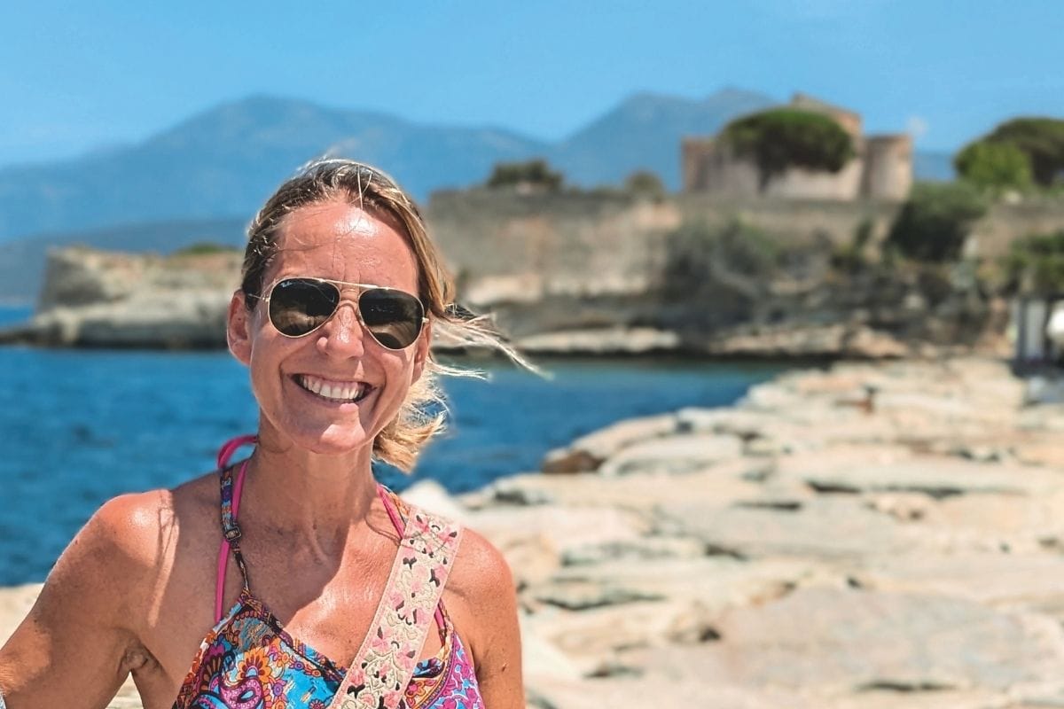 Eine lächelnde Frau mit Sonnenbrille steht vor einer Küstenlandschaft mit blauem Meer, Felsen und einer historischen Festungsanlage im Hintergrund.