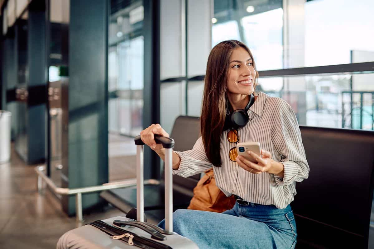 Eine lächelnde Frau mit langem braunem Haar sitzt in einem modernen Terminal, hält ein Smartphone in der Hand und greift nach dem Griff ihres Rollkoffers.