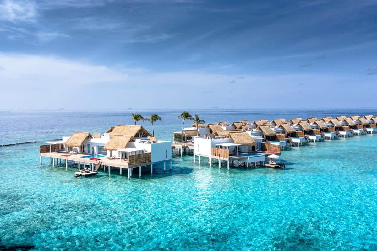 Eine Reihe von weißen Villen mit Strohdächern steht auf Stelzen über dem türkisfarbenen Wasser eines tropischen Ozeans unter einem klaren, blauen Himmel.