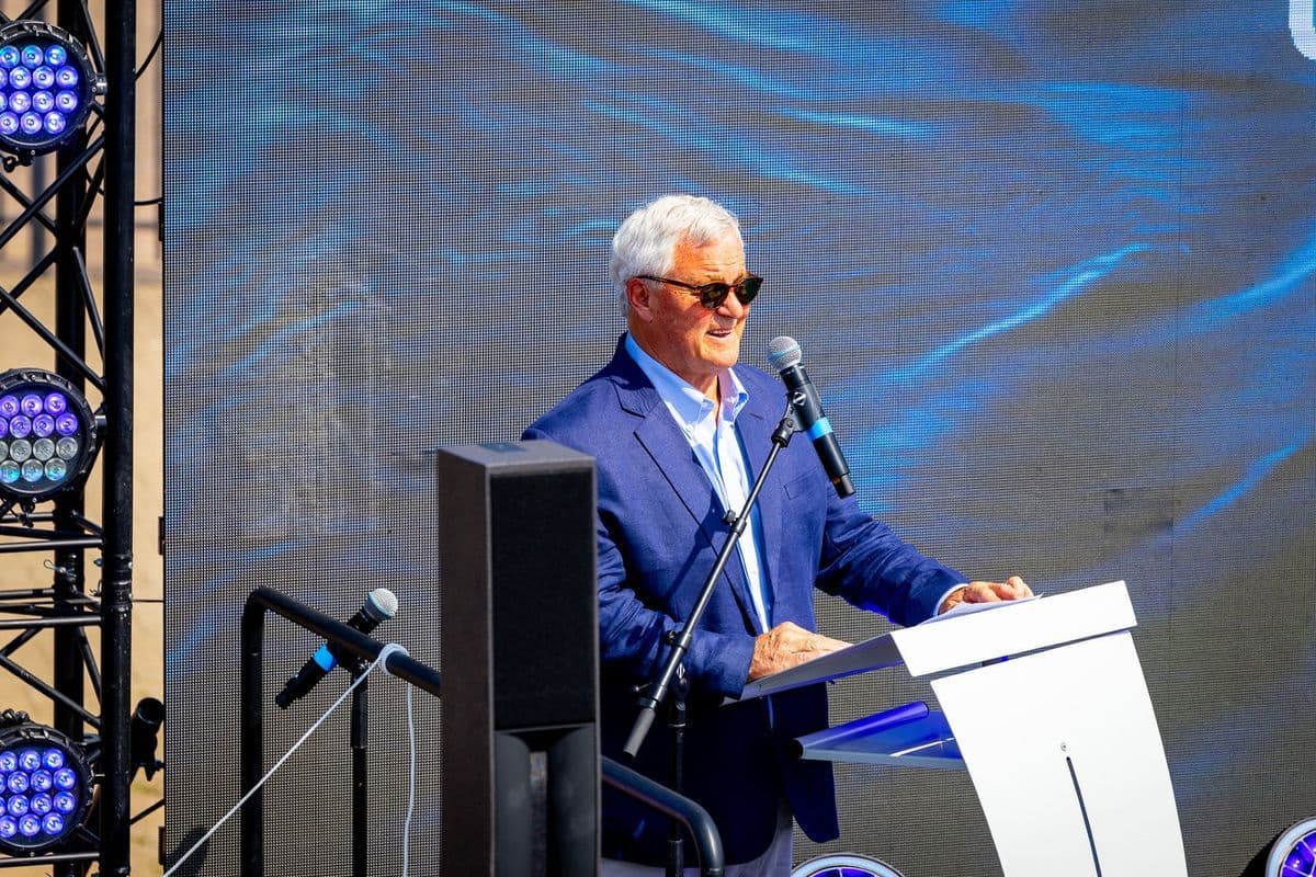 Ein älterer Mann mit grauem Haar und Sonnenbrille trägt ein blaues Sakko und spricht an einem Rednerpult in ein Mikrofon vor einer großen LED-Bildwand.