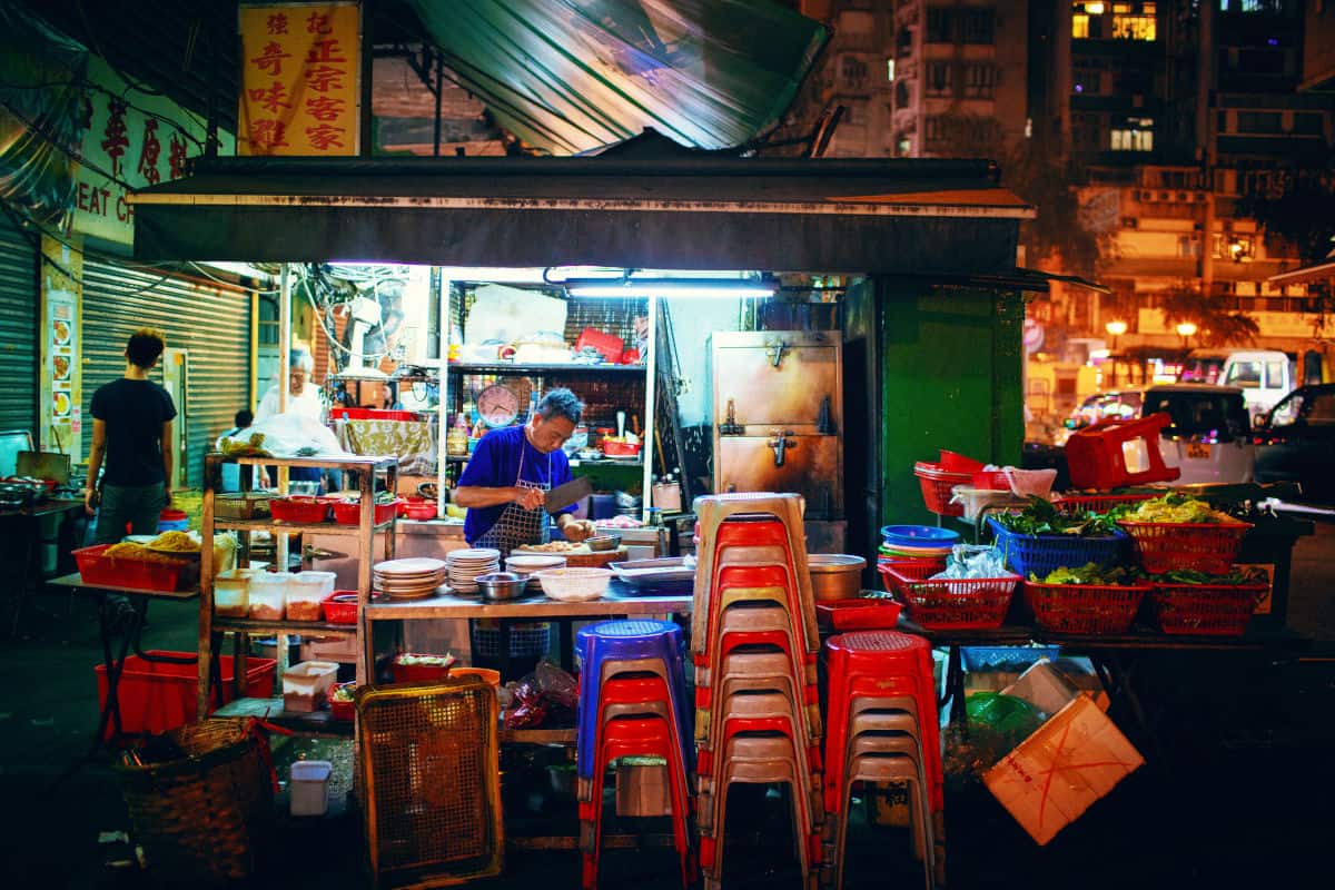 Ein Mann bereitet in einem beleuchteten Straßenimbiss in Hongkong Speisen zu, davor stehen gestapelte bunte Kunststoffhocker auf dem nächtlichen Gehweg.