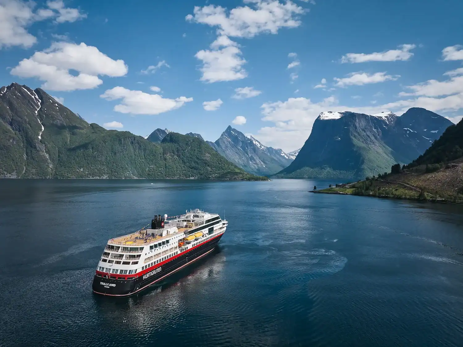Das Hurtigruten-Schiff Trollfjord fährt durch einen tiefblauen norwegischen Fjord, umgeben von hohen, teils schneebedeckten Bergen unter einem sonnigen Himmel.