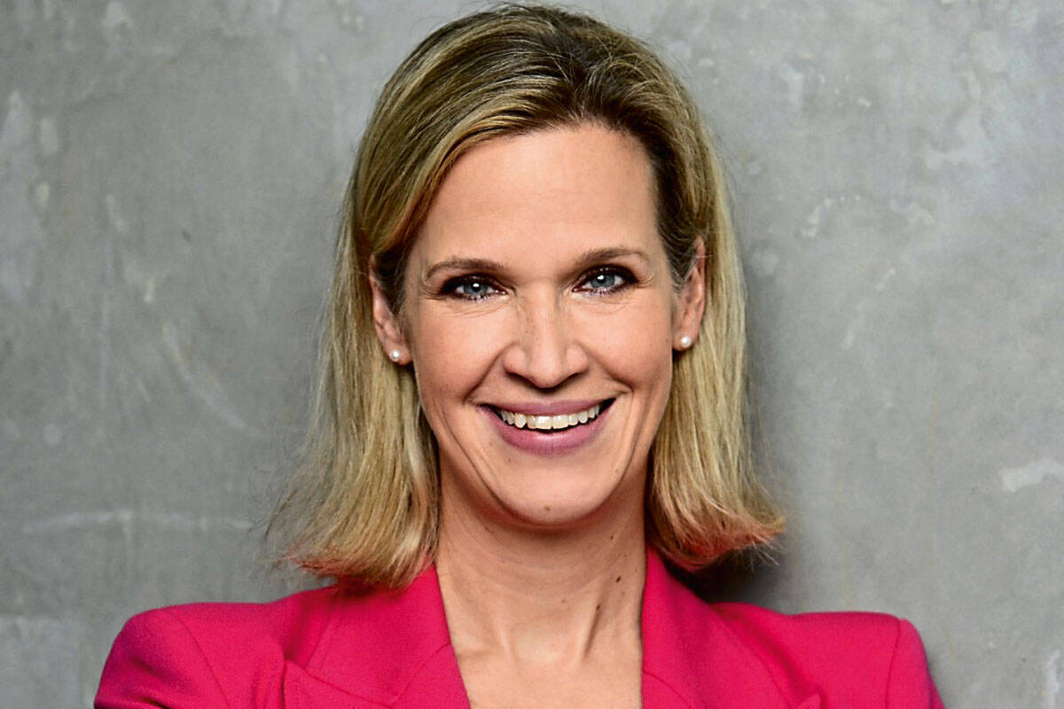 Eine blonde Frau mit schulterlangem Haar und Perlenohrringen lächelt freundlich in die Kamera. Sie trägt einen leuchtend pinken Blazer vor grauer Wand.
