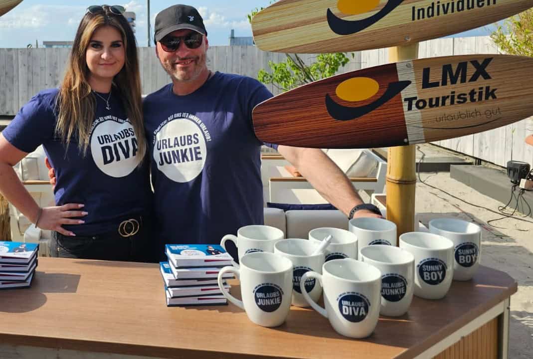Eine Frau und ein Mann stehen hinter einem Tresen mit Tassen und Broschüren, daneben ein Pfahl mit Schildern in Surfbrettoptik und dem Logo LMX Touristik.