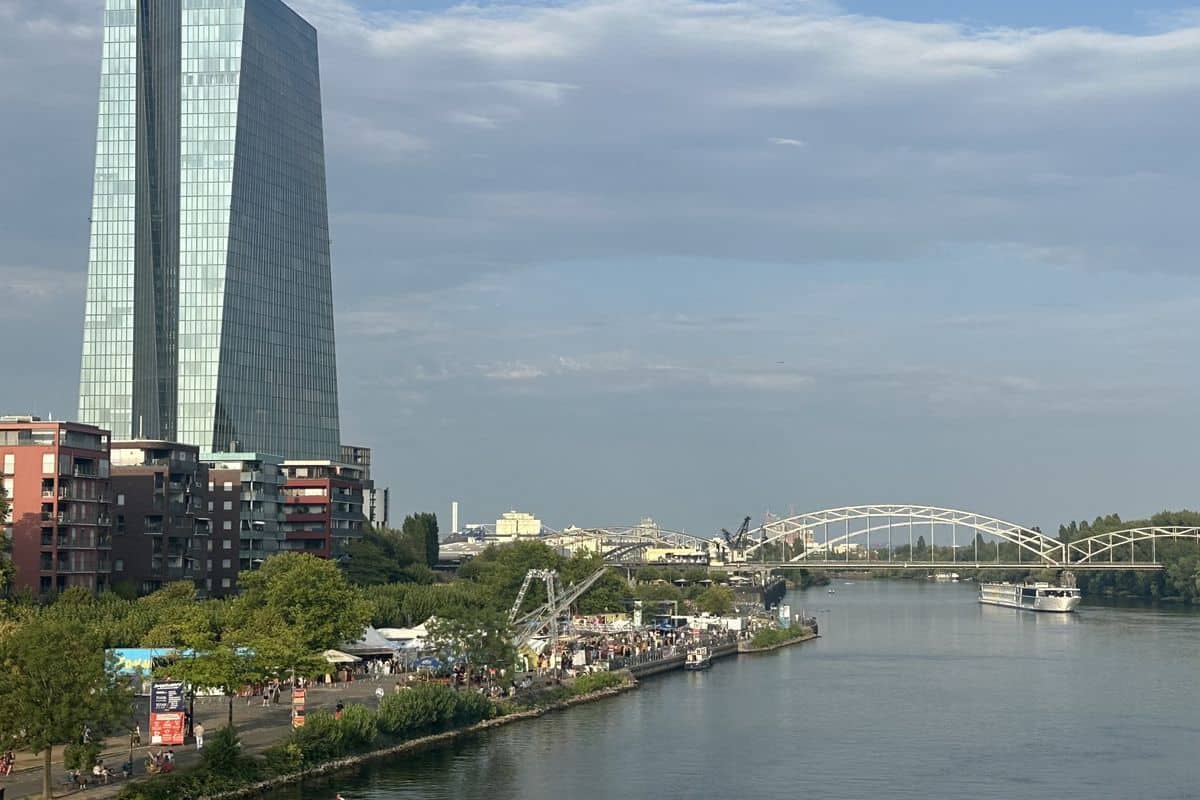 Blick auf den Main in Frankfurt mit dem EZB-Hochhaus am linken Ufer, einer Bogenbrücke im Hintergrund und einem Flusskreuzfahrtschiff auf dem Wasser.