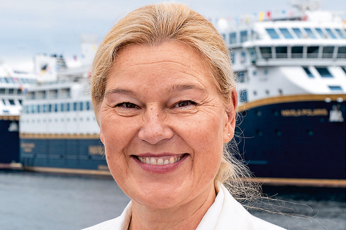 Eine blonde Frau mittleren Alters lächelt freundlich in die Kamera, im Hintergrund ist ein großes Kreuzfahrtschiff an einem Hafenbecken zu sehen.