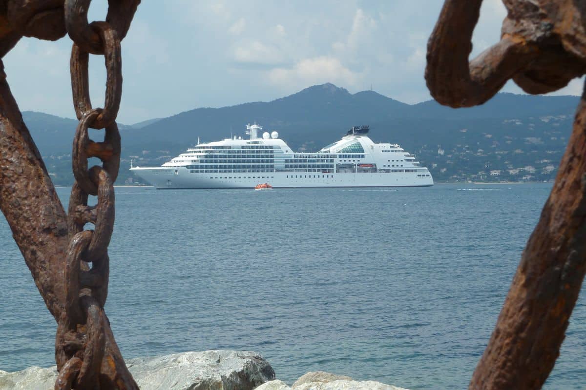 Ein weißes Kreuzfahrtschiff liegt auf dem blauen Meer vor einer hügeligen Küste, eingerahmt von den rostigen, massiven Gliedern einer großen Ankerkette.