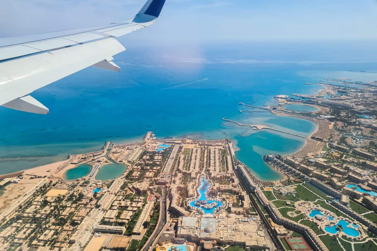 Blick aus einem Flugzeugfenster auf eine Küstenregion mit Hotelanlagen, Swimmingpools und Sandstränden, die an das tiefblaue Meer grenzen, bei klarem Himmel.