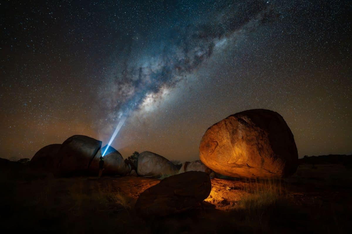 Eine Person steht nachts zwischen großen Felsblöcken in einer Wüstenlandschaft und richtet eine Taschenlampe mit einem Lichtstrahl in den Sternenhimmel.