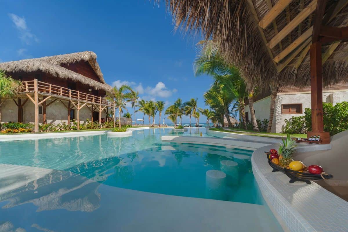 Ein tropischer Hotelpool mit Swim-up-Bar, umgeben von strohgedeckten Gebäuden, Palmen und einer Obstschale am Beckenrand unter einem strahlend blauen Himmel.