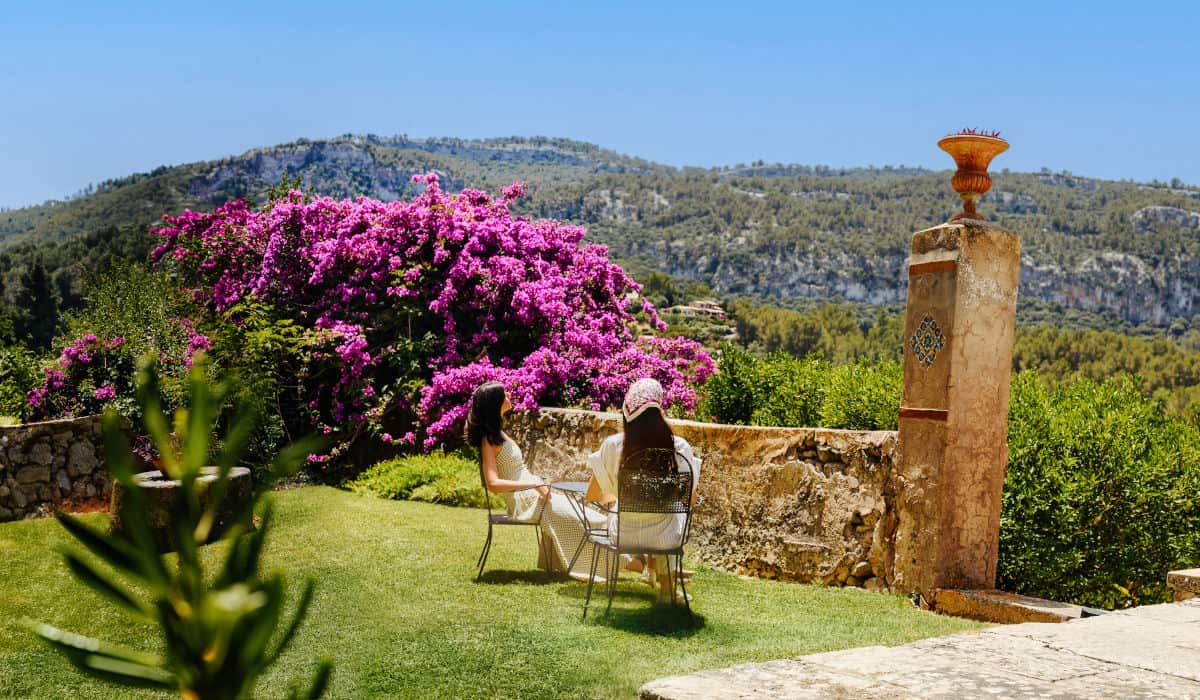 Zwei Frauen sitzen auf Stühlen in einem sonnigen Garten vor einer großen, violett blühenden Bougainvillea mit Blick auf eine hügelige, bewaldete Landschaft.