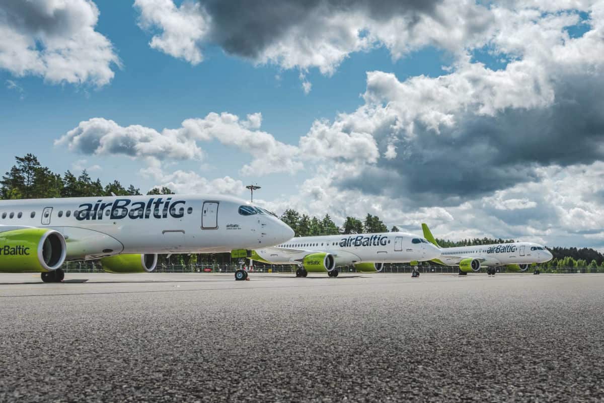 Drei weiße Passagierflugzeuge der Fluggesellschaft airBaltic stehen hintereinander auf einem Rollfeld vor einem bewaldeten Hintergrund bei bewölktem Himmel.