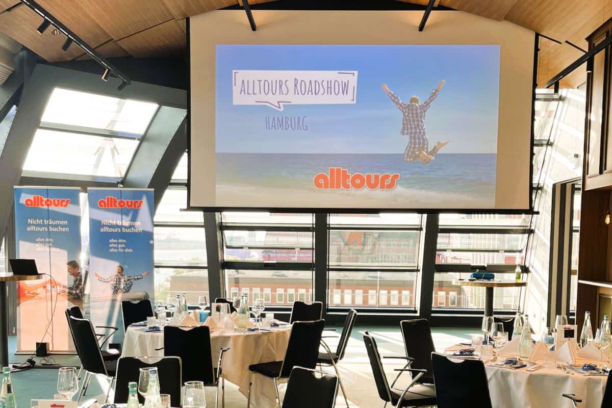 Eine Leinwand mit der Aufschrift Alltours Roadshow Hamburg und ein Bild eines springenden Menschen über dem Meer, flankiert von zwei Alltours-Roll-ups.