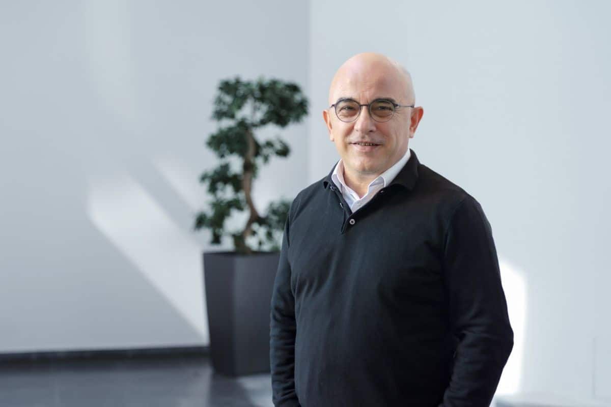 Ein kahlköpfiger Mann mit Brille trägt einen schwarzen Pullover über einem weißen Hemd und lächelt vor einer hellen Wand mit einer Topfpflanze im Hintergrund.