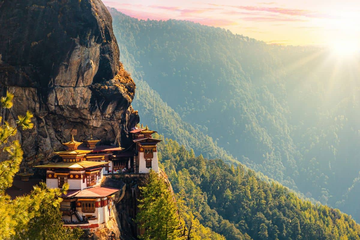 Das Kloster Taktshang in Bhutan schmiegt sich an eine steile Felswand über einem bewaldeten Tal, während die Sonne im Hintergrund sanft hinter Bergen aufgeht.