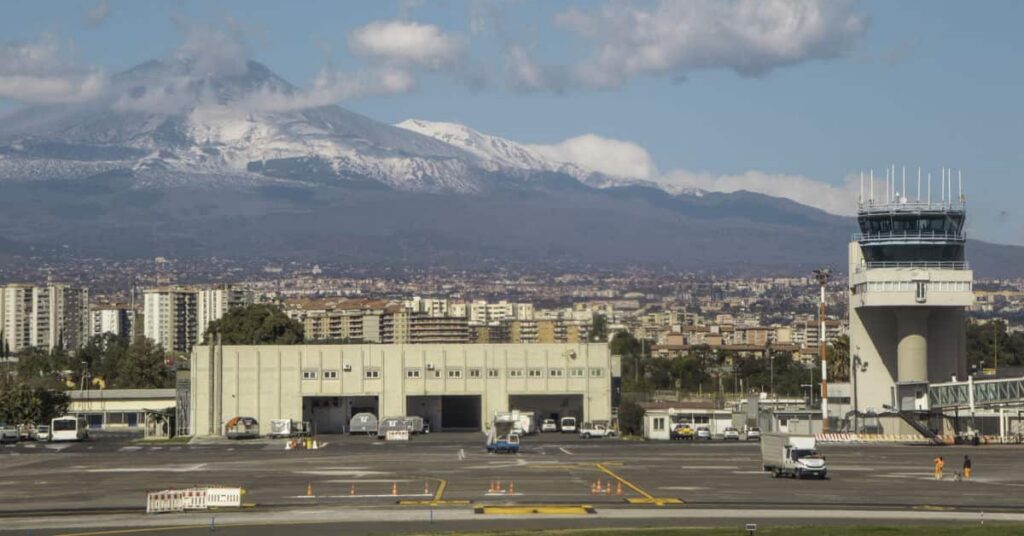 Blick auf das Flughafengelände mit einem Kontrollturm, im Hintergrund erstreckt sich die Stadt Catania vor dem schneebedeckten Gipfel des Ätna unter blauem Himmel.
