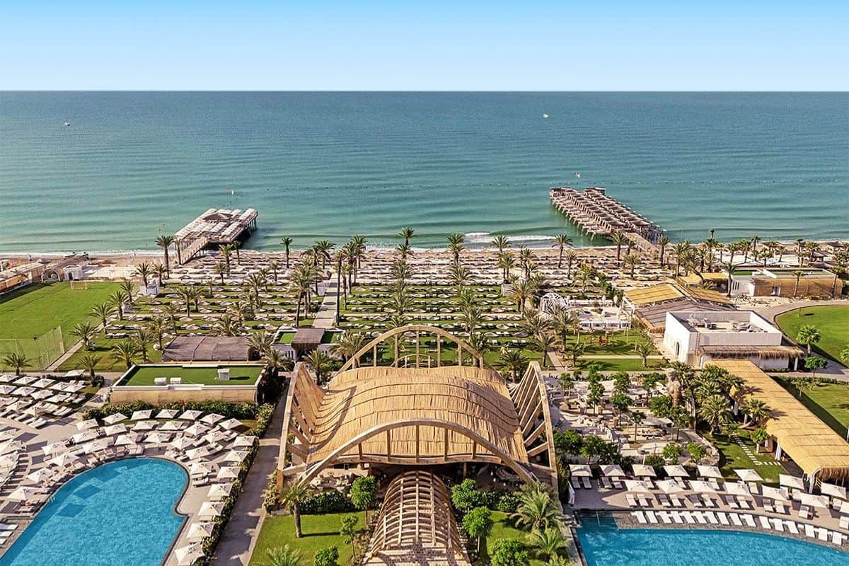Luftaufnahme einer Hotelanlage mit zwei Pools, einer markanten Holzkonstruktion, zahlreichen Palmen, Liegen am Strand und zwei Stegen im türkisfarbenen Meer.
