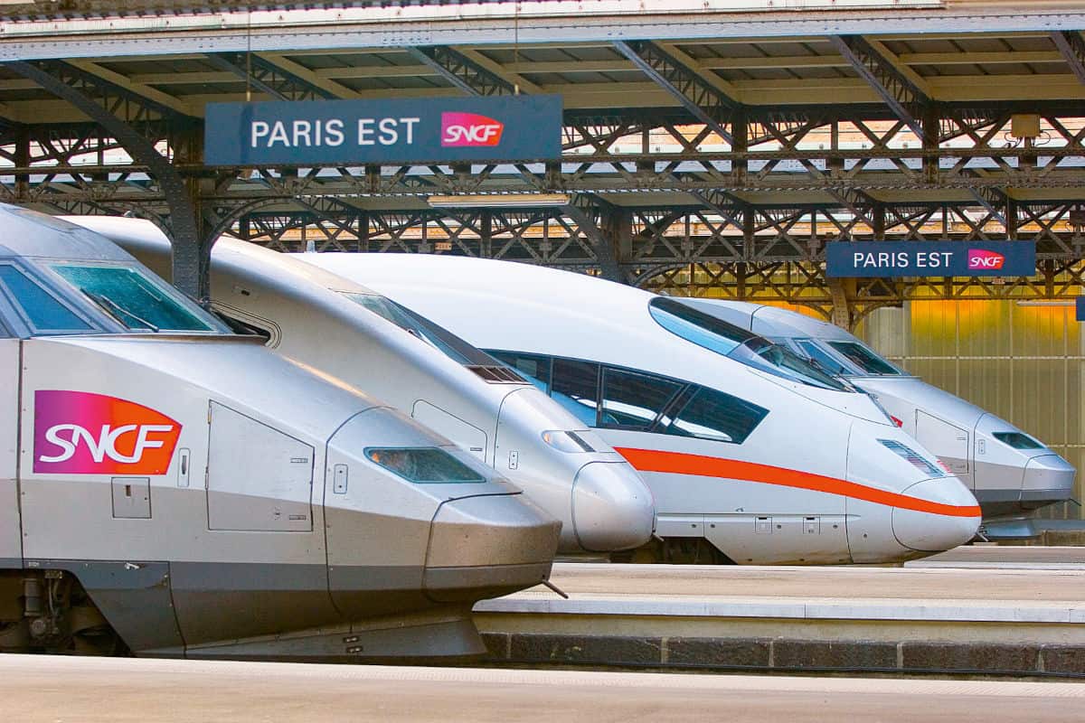 Mehrere silberne Hochgeschwindigkeitszüge stehen nebeneinander an den Bahnsteigen des Bahnhofs Paris Est unter einer großen, historischen Stahlkonstruktion.