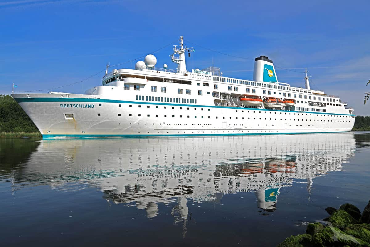 Das weiße Kreuzfahrtschiff Deutschland fährt auf einem ruhigen Gewässer vor einem klaren blauen Himmel, wobei sich das Schiff deutlich im Wasser spiegelt.