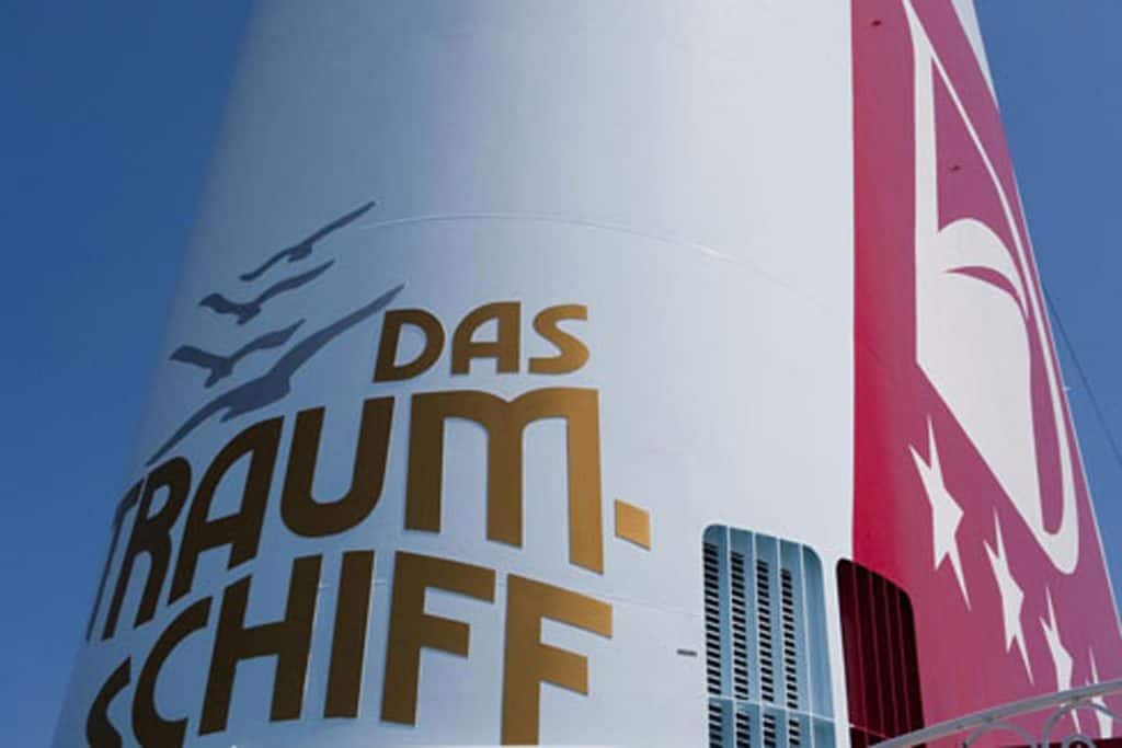 Nahaufnahme des weißen Schornsteins eines Kreuzfahrtschiffes mit dem goldenen Schriftzug Das Traumschiff und einer roten Fläche mit weißen Sternen.