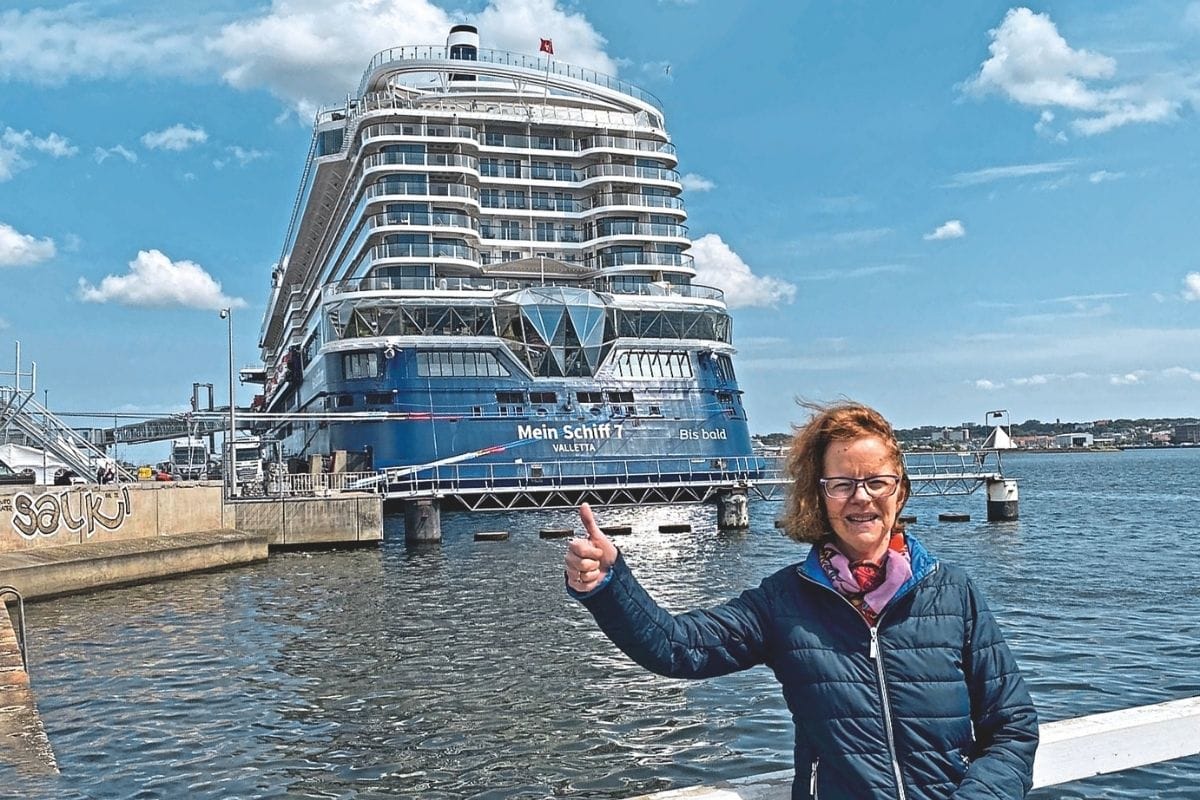 Eine Frau mit Brille und dunkelblauer Steppjacke steht im Vordergrund an einem Hafen und zeigt mit dem Daumen nach oben vor dem Kreuzfahrtschiff Mein Schiff 7.
