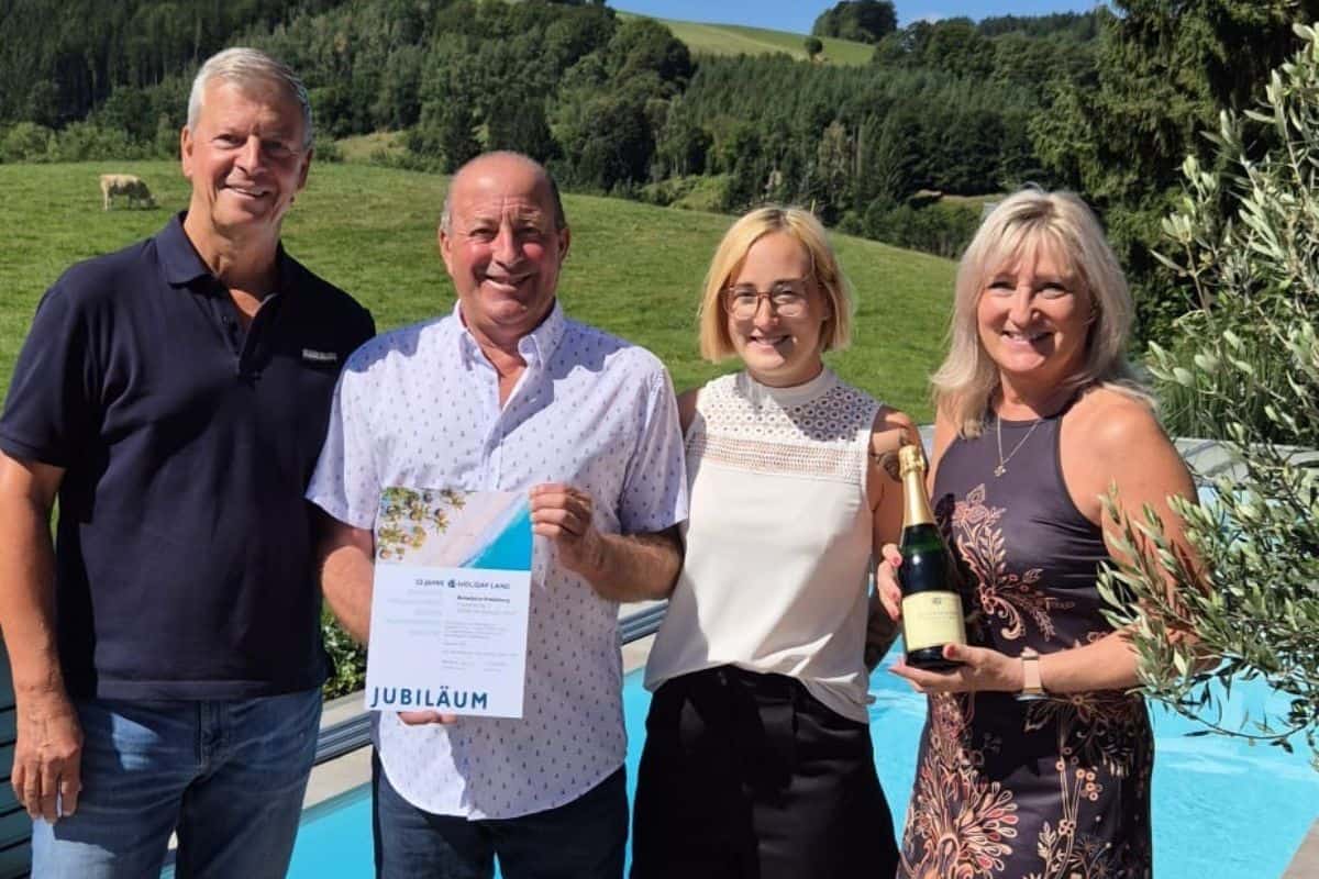 Zwei Männer und zwei Frauen stehen lächelnd vor einem Pool in einer grünen Hügellandschaft. Einer hält eine Urkunde, die andere eine Flasche Sekt in Händen.