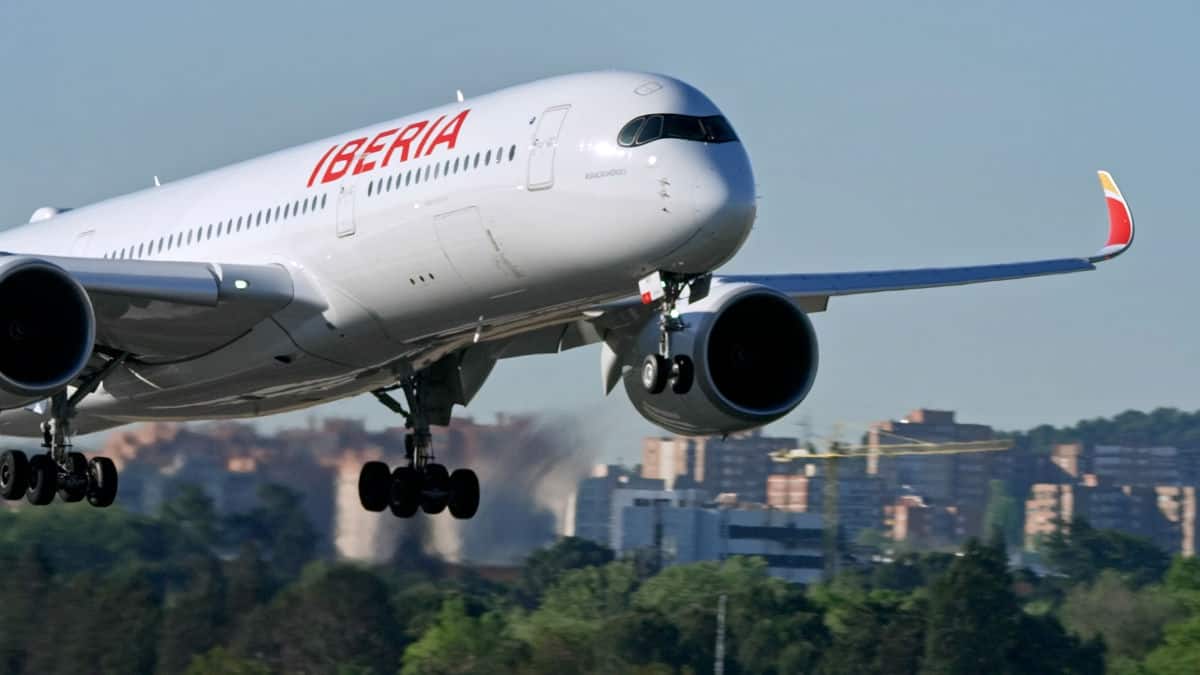 Ein weißes Passagierflugzeug der Fluggesellschaft Iberia im Landeanflug mit ausgefahrenem Fahrwerk vor einer städtischen Kulisse mit Bäumen und Gebäuden.