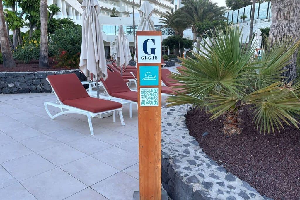 Ein hölzerner Pfosten mit Beschilderung für Liegenbereiche steht auf einer Terrasse vor einer Reihe roter Sonnenliegen und geschlossenen Sonnenschirmen.