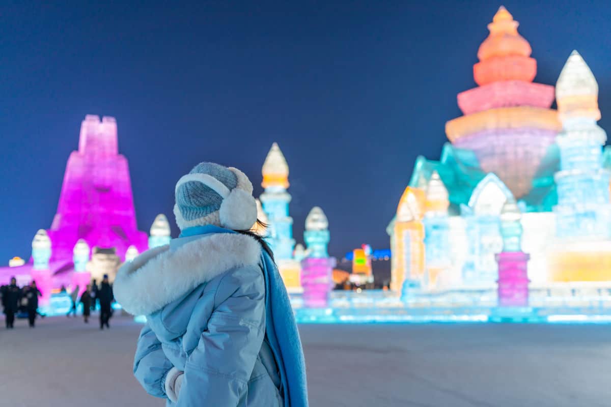 Eine Person in Winterkleidung betrachtet bei Nacht beleuchtete, farbenfrohe Eisskulpturen und Gebäude aus Eis auf einem weitläufigen, verschneiten Gelände.
