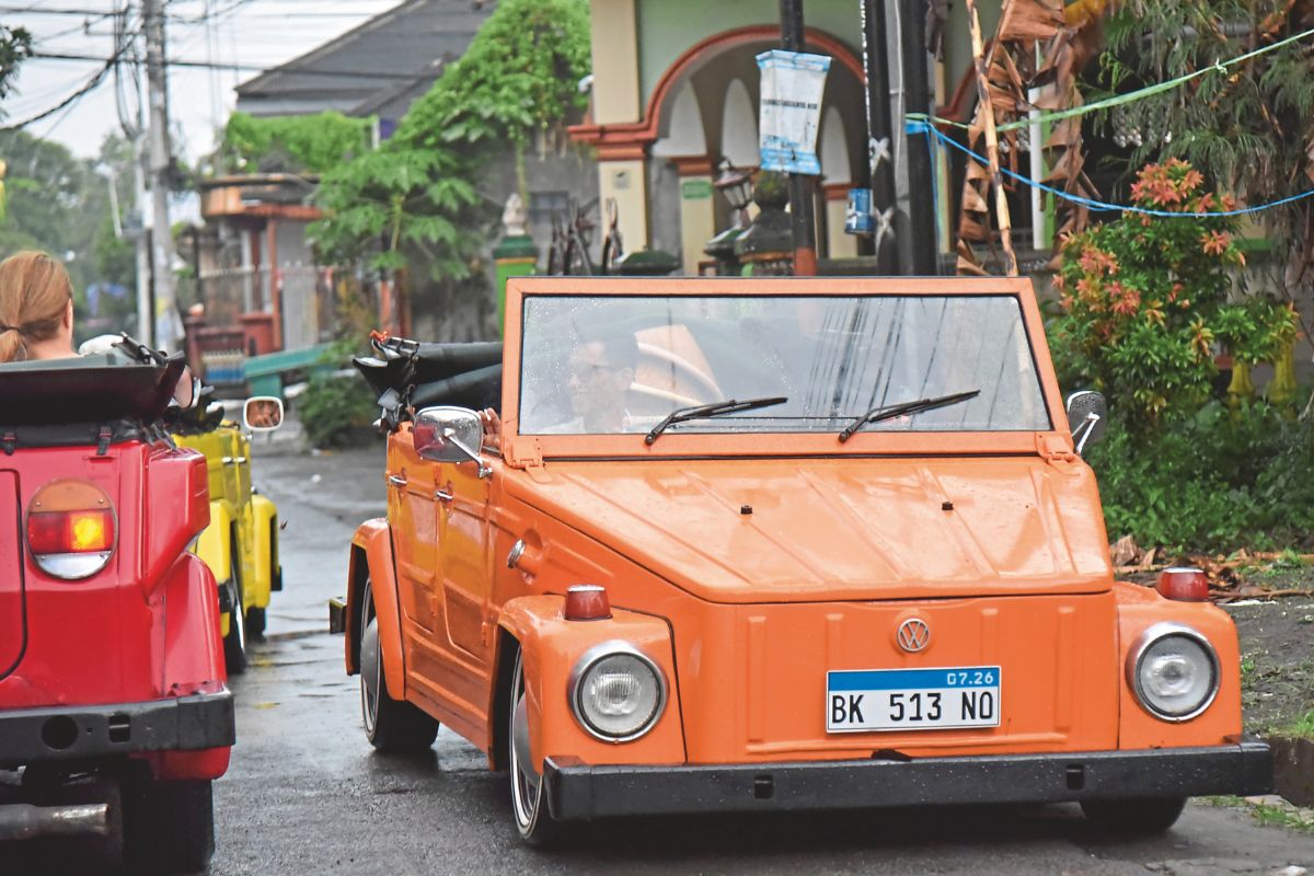 Ein orangefarbener Volkswagen Kübelwagen fährt auf einer Straße, gefolgt von einem gelben und einem roten Fahrzeug derselben Bauart in einer Wohngegend.