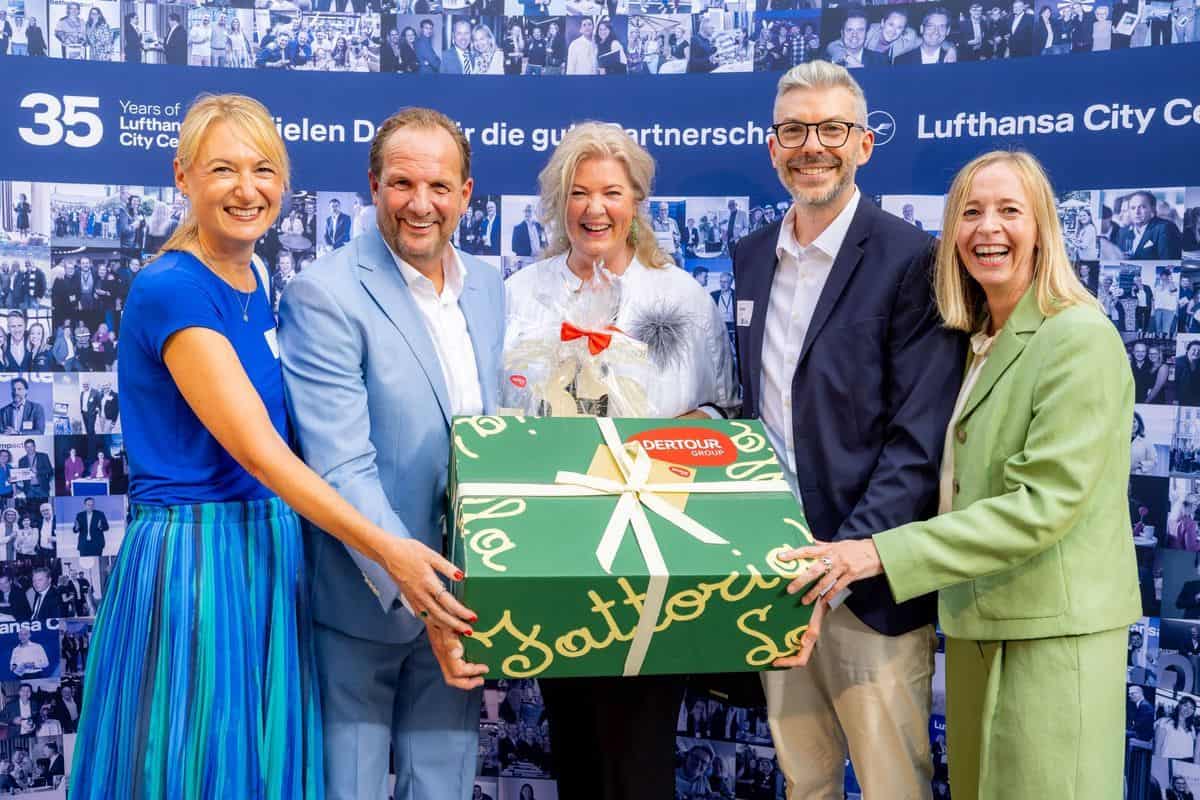 Fünf Personen stehen vor einer Fotowand und halten gemeinsam ein großes, grün verpacktes Geschenk mit weißer Schleife und Dertour Group Logo in den Händen.