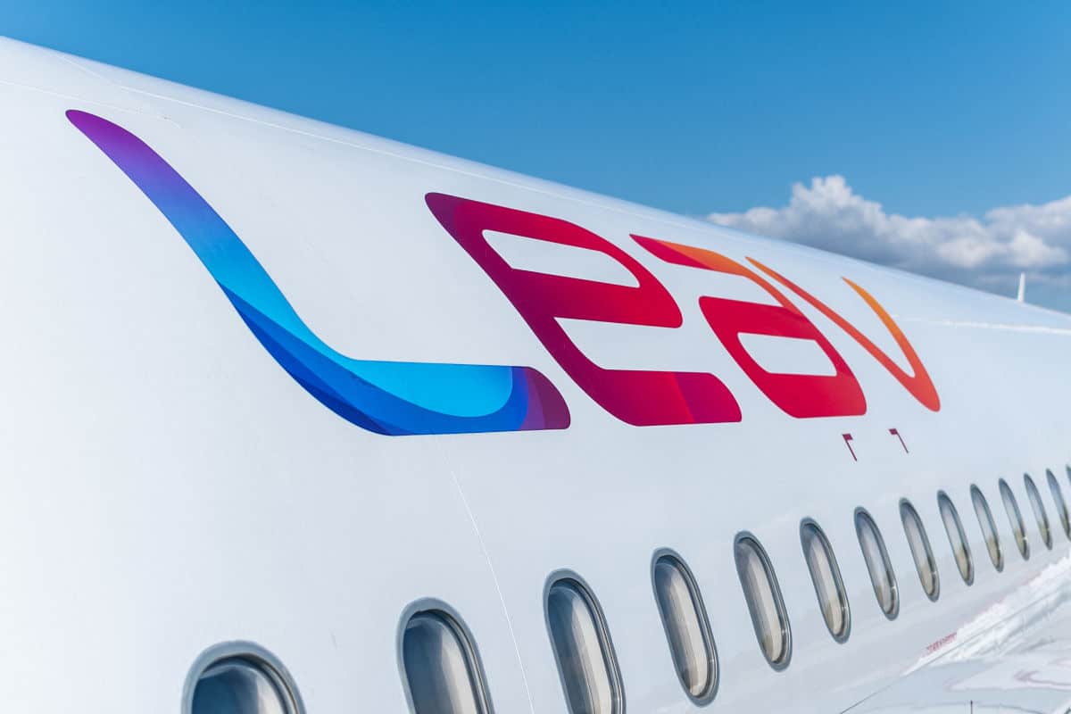 Nahaufnahme der weißen Außenhülle eines Flugzeugs mit dem farbigen LEAV-Schriftzug und einer Reihe von ovalen Kabinenfenstern vor einem blauen Himmel.