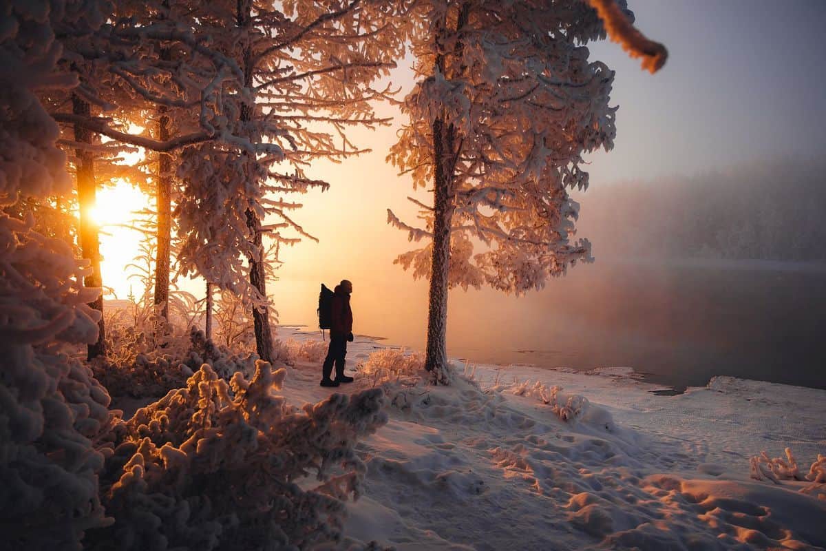 Eine Person mit Rucksack steht im Profil am Ufer eines verschneiten Flusses, umgeben von frostbedeckten Bäumen im warmen Licht eines tiefstehenden Sonnenaufgangs.