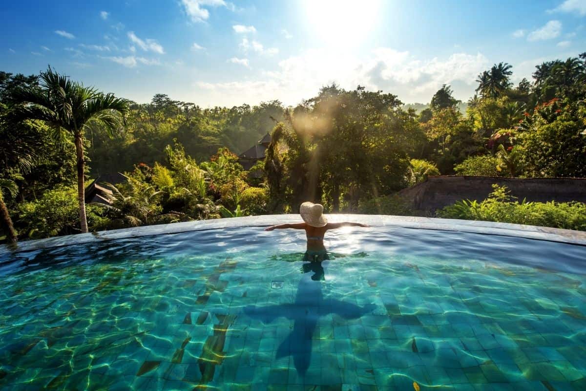 Eine Person mit Sonnenhut steht mit ausgebreiteten Armen in einem Infinity-Pool vor einer üppigen tropischen Waldlandschaft im hellen Gegenlicht der Sonne.