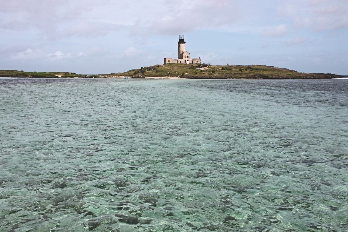 Ein einsamer Leuchtturm steht auf einer flachen, grasbewachsenen Insel inmitten von klarem, türkisblauem Wasser unter einem bewölkten Himmel bei Tageslicht.