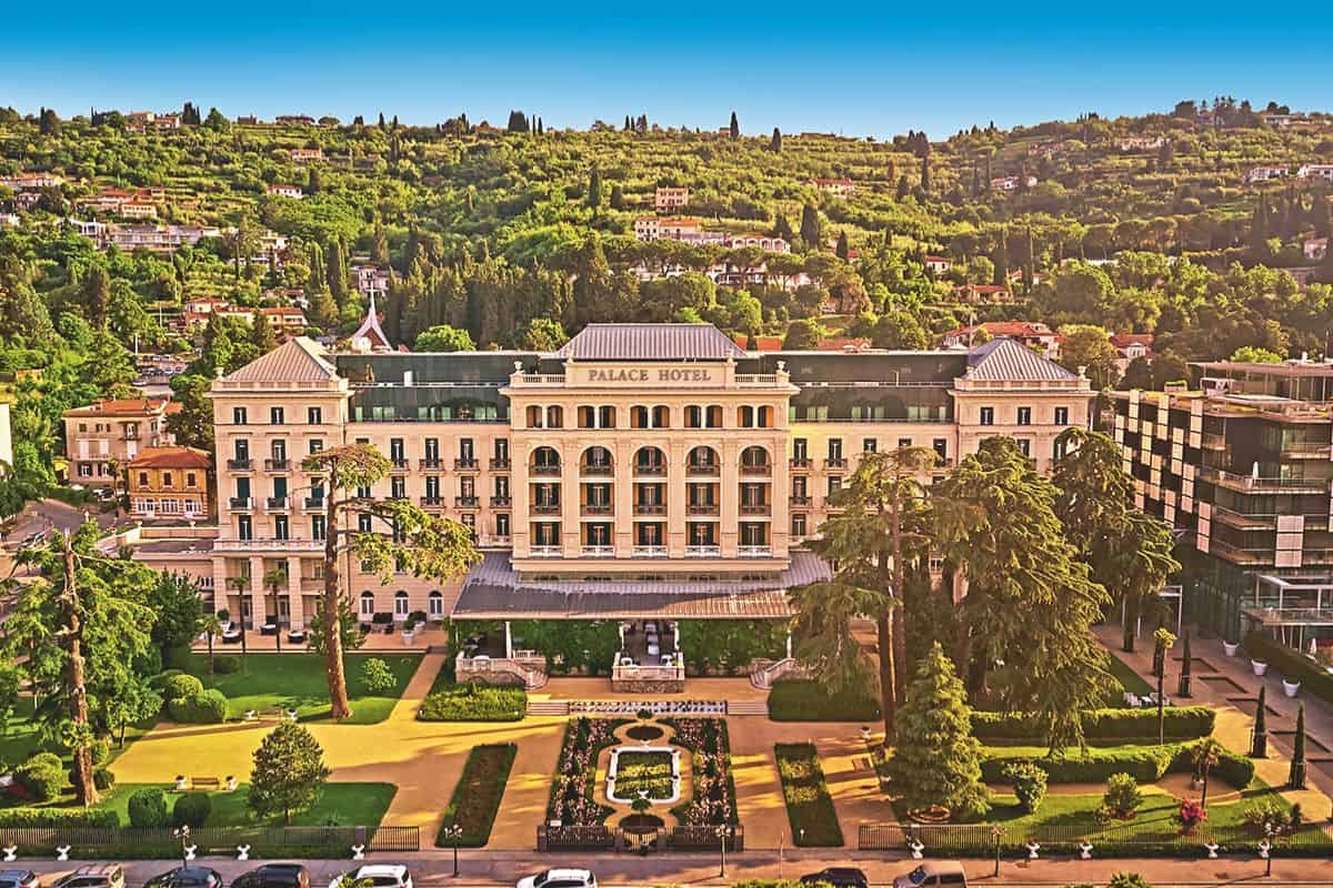 Eine Frontalaufnahme des Palace Hotel, umgeben von einer weitläufigen Gartenanlage mit Blumenbeeten und Bäumen vor einem bewaldeten Hügel bei Tageslicht.