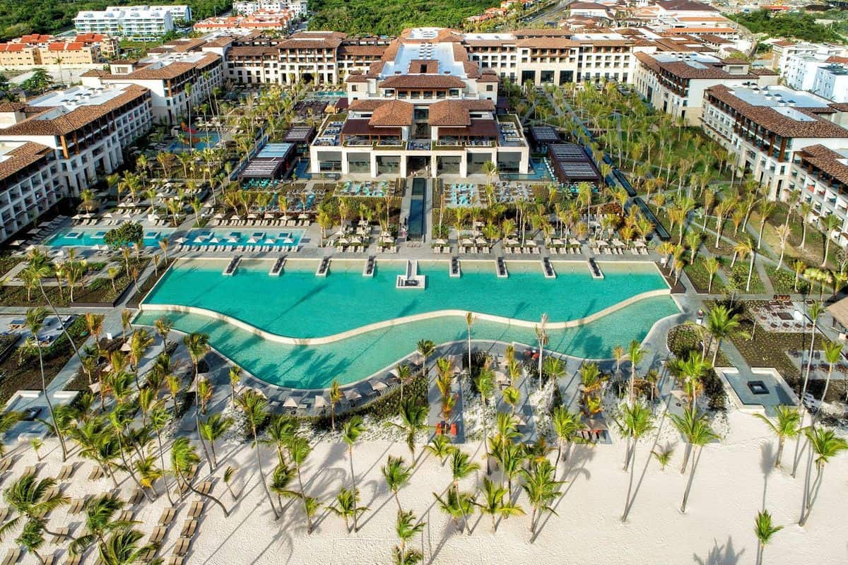 Luftaufnahme eines weitläufigen Hotelresorts mit großen Swimmingpools, Palmen, Sandstrand und Gebäuden mit Terrakottadächern in einer tropischen Umgebung.