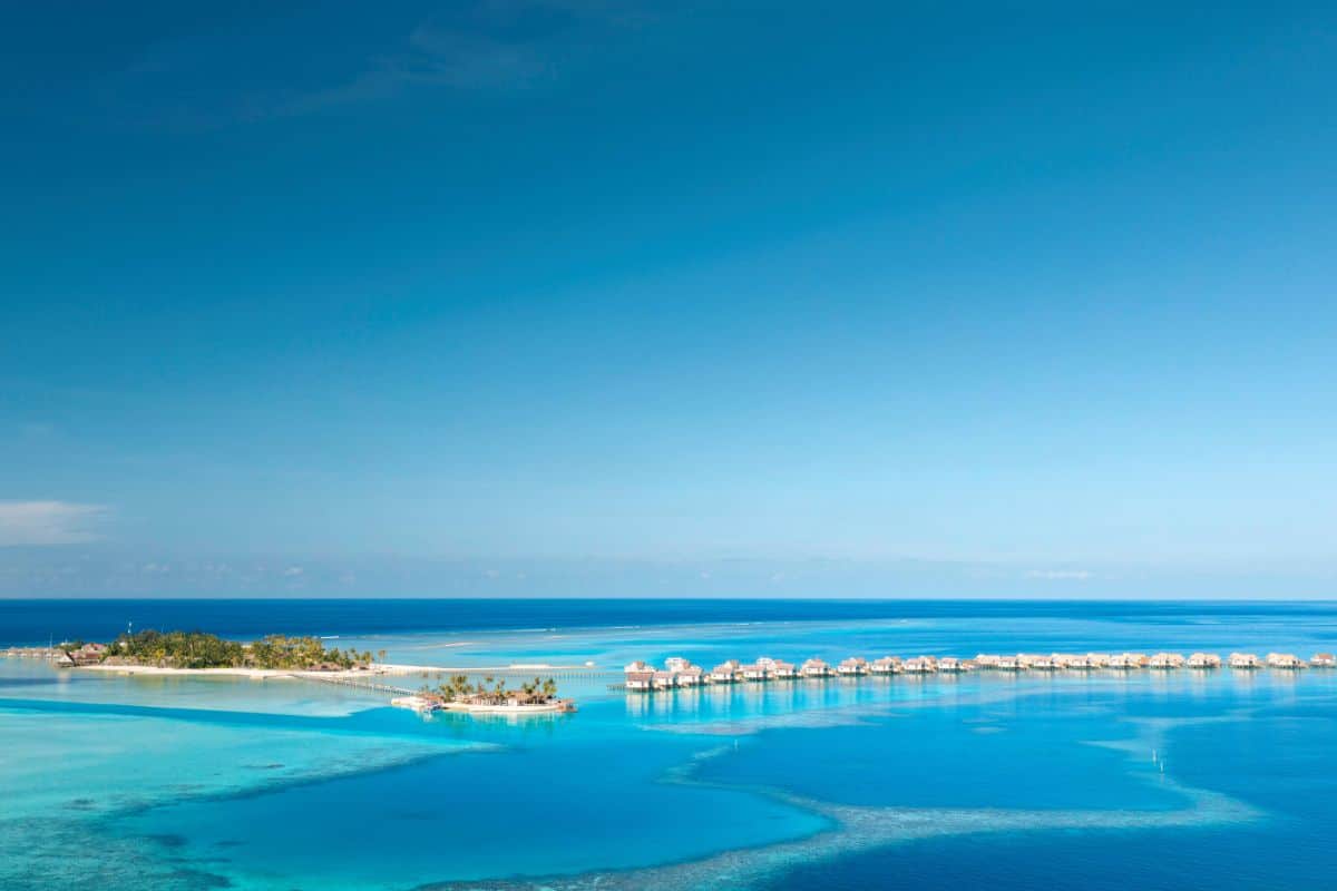 Luftaufnahme einer tropischen Inselgruppe mit zahlreichen über dem türkisfarbenen Wasser errichteten Bungalows unter einem klaren, tiefblauen Himmel.