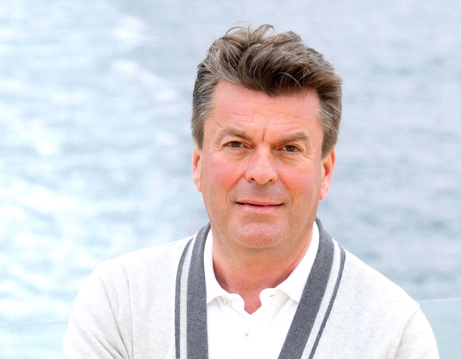 Ein Mann mit braunem Haar und freundlichem Gesichtsausdruck trägt eine weiße Strickjacke mit grauen Streifen vor einem unscharfen, hellblauen Wasserhintergrund.