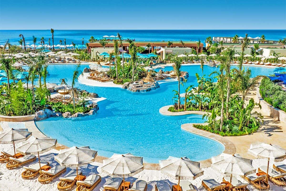 Eine weitläufige Hotel-Poolanlage mit türkisblauem Wasser, zahlreichen Palmen, hellen Sonnenschirmen und Liegen vor einem tiefblauen Meer im Hintergrund.
