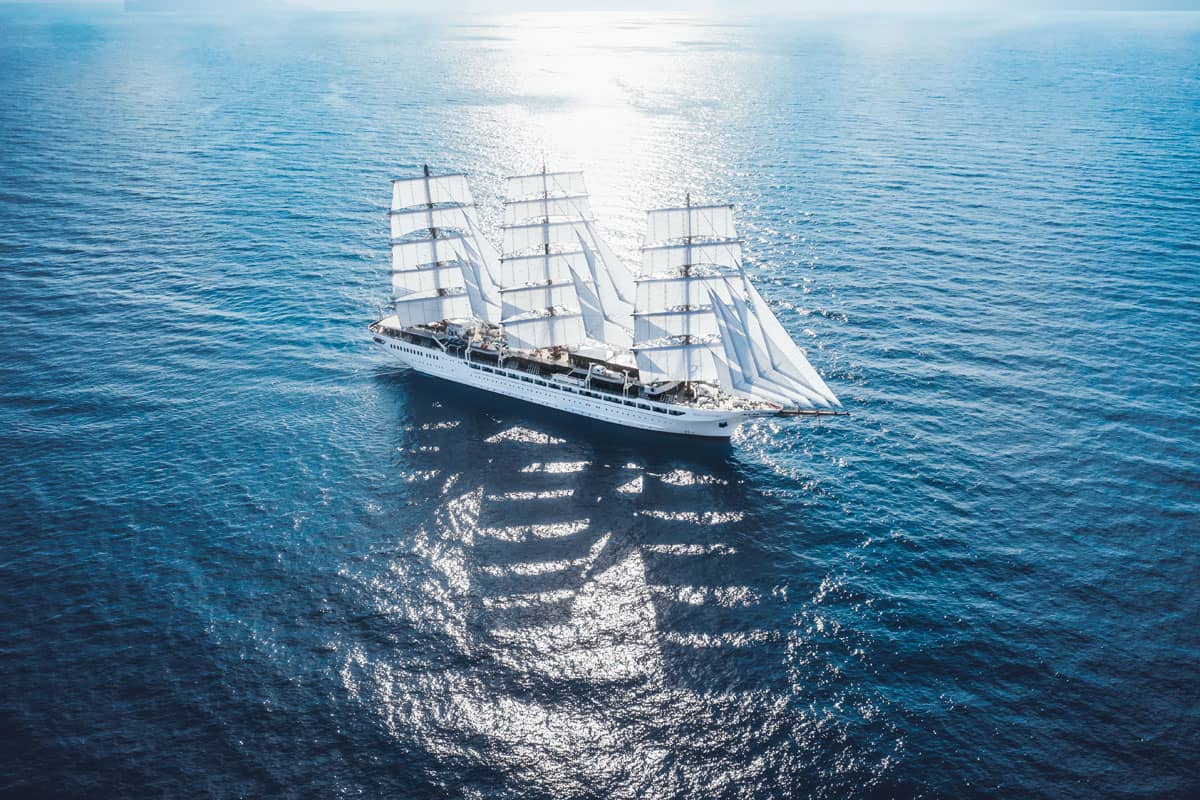 Eine Luftaufnahme eines großen weißen Segelschiffs mit fünf Masten, das bei hellem Sonnenlicht auf dem tiefblauen, leicht bewegten Wasser des Meeres fährt.